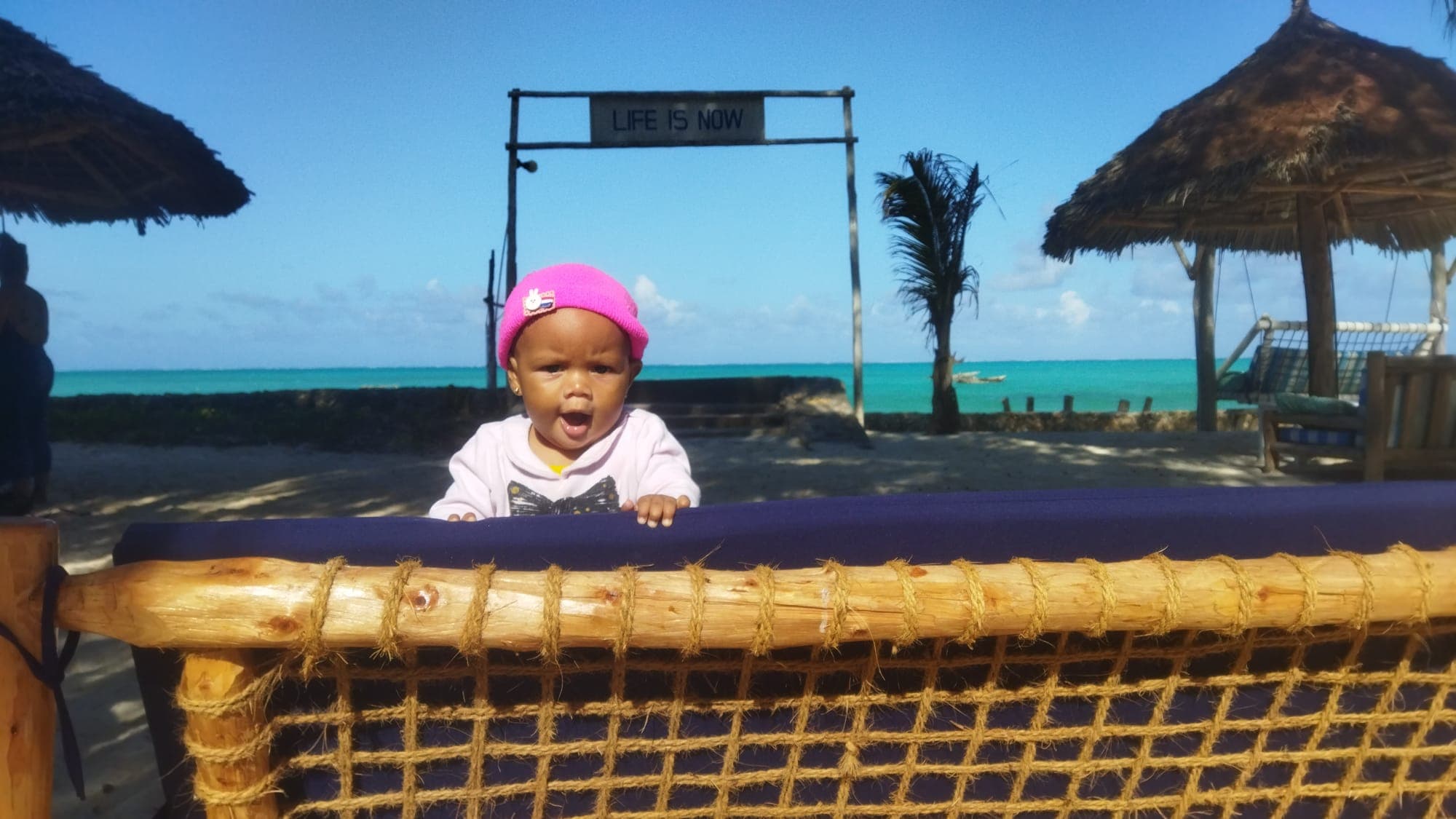 A baby at a beach on Zanzibar, thatched sun shades and turquoise water behind a 'Life Is Now' sign