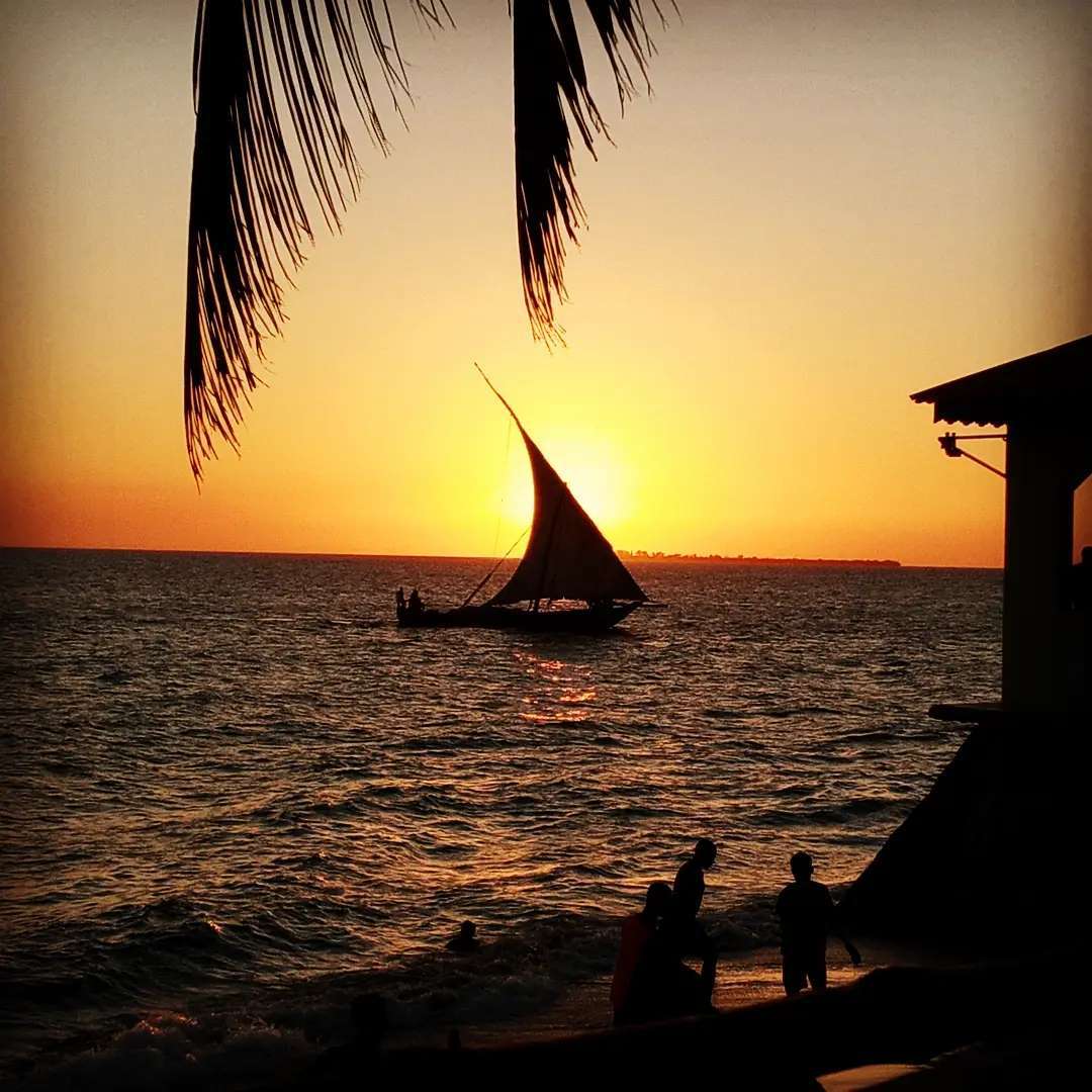 A traditional Dhow boat sailing home at sunset off Zanzibar's Swahili Coast, families gathered at the water's edge